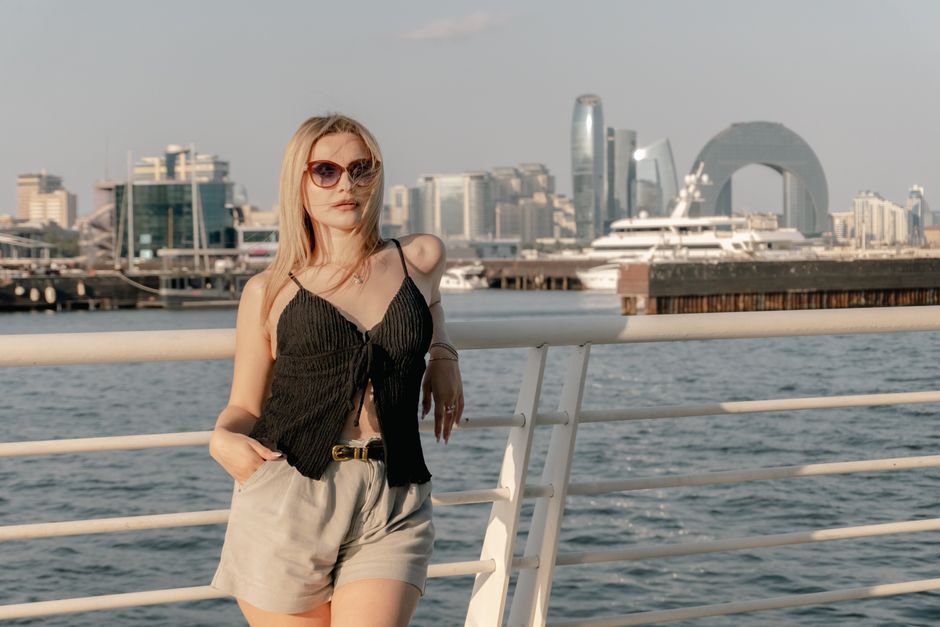 Woman in chic summer attire posing by a waterfront with a modern cityscape backdrop.