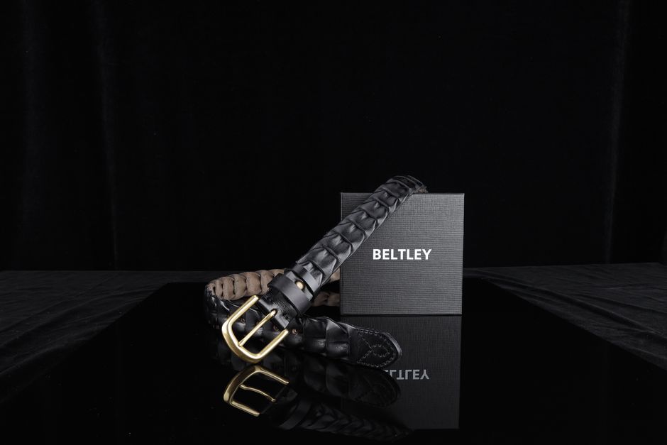 Elegant black leather belt with gold buckle displayed on reflective surface with branded packaging.
