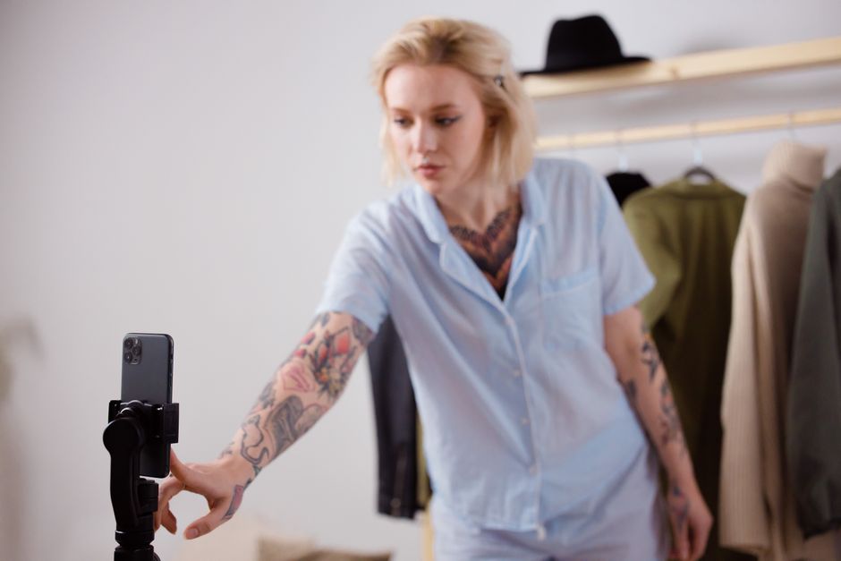A stylish woman with tattoos adjusting her smartphone on a tripod, surrounded by clothing.