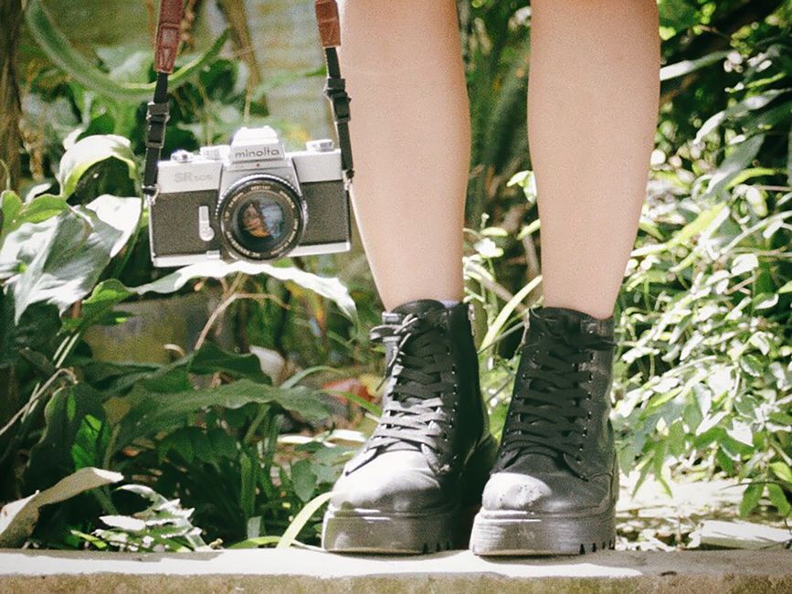 Legs with black boots and vintage camera in a lush garden setting.