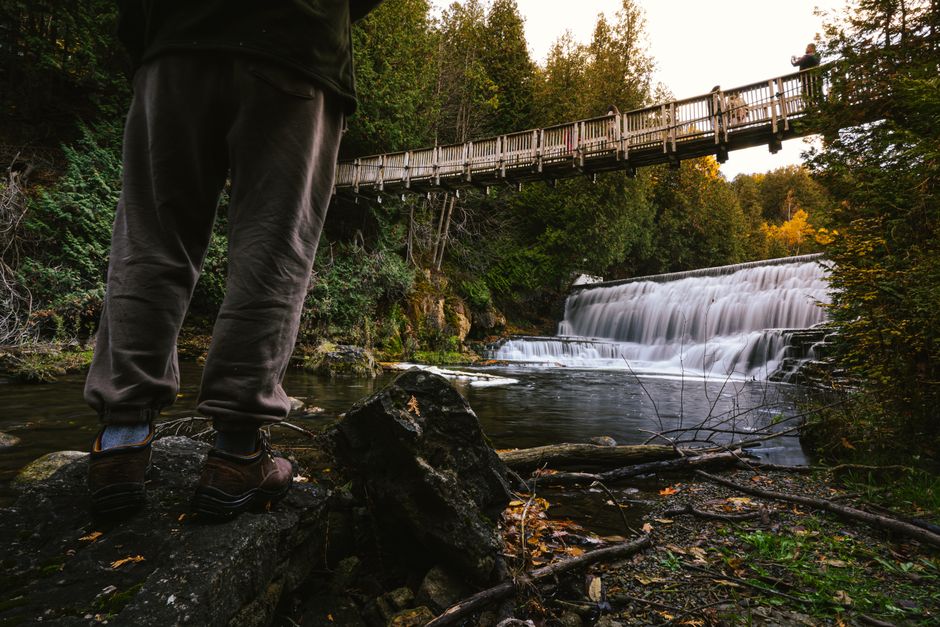 A person stands near a waterfall and bridge, capturing nature's beauty on an adventure.