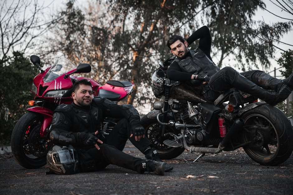 Two bikers in leather gear pose with motorcycles on an outdoor road.