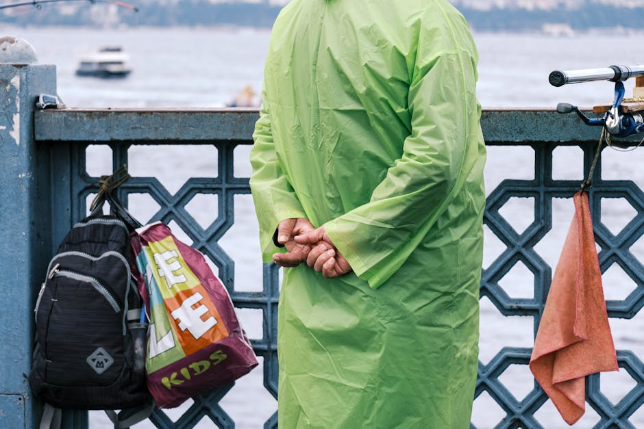 A man in a neon green raincoat stands on a waterfront bridge with bags and a towel.