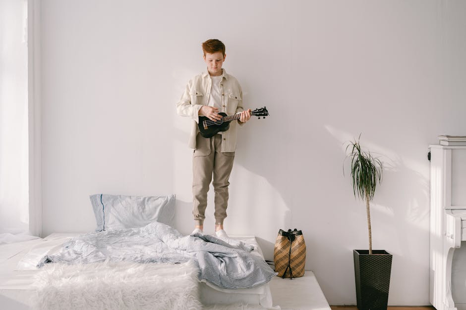 Teen boy plays ukulele while standing on a bed in a bright and airy bedroom setting.