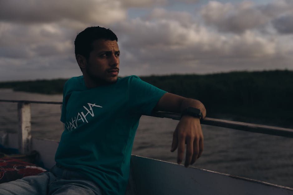 A man in a teal shirt relaxes on a boat during a cloudy evening, evoking a contemplative mood.