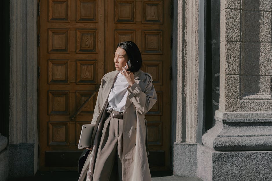 Fashionable Asian businesswoman in a trench coat making a phone call outside a wooden door.
