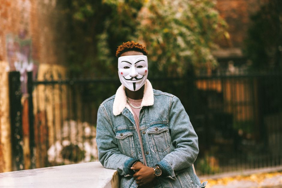 Anonymous African American male with dyed hair standing with clasped hands in mask of criminal comic character in autumn town