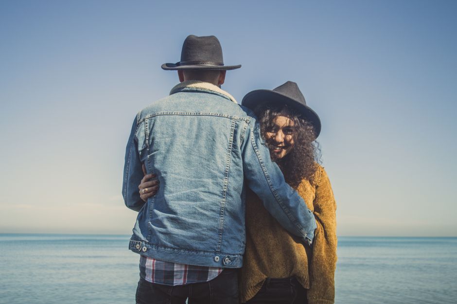 A couple embraces at the seaside wearing casual outfits and hats, enjoying a sunny day.