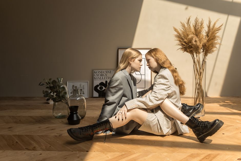 Two women in stylish outfits sit on a wooden floor in a sunlit room with decorative vases.