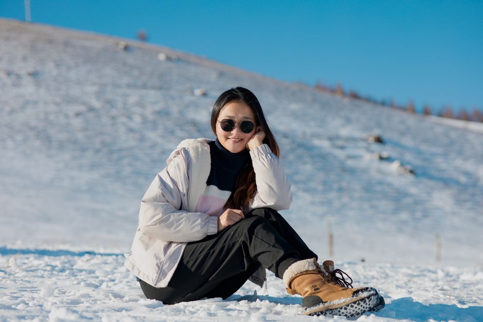 Smiling young woman sitting in the snow wearing sunglasses and warm clothing on a sunny winter day.