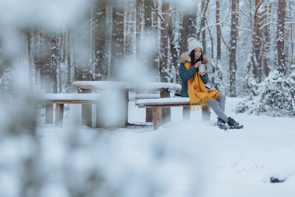 Asian woman in winter attire sipping a drink in a snowy forest. Serene and cozy atmosphere.