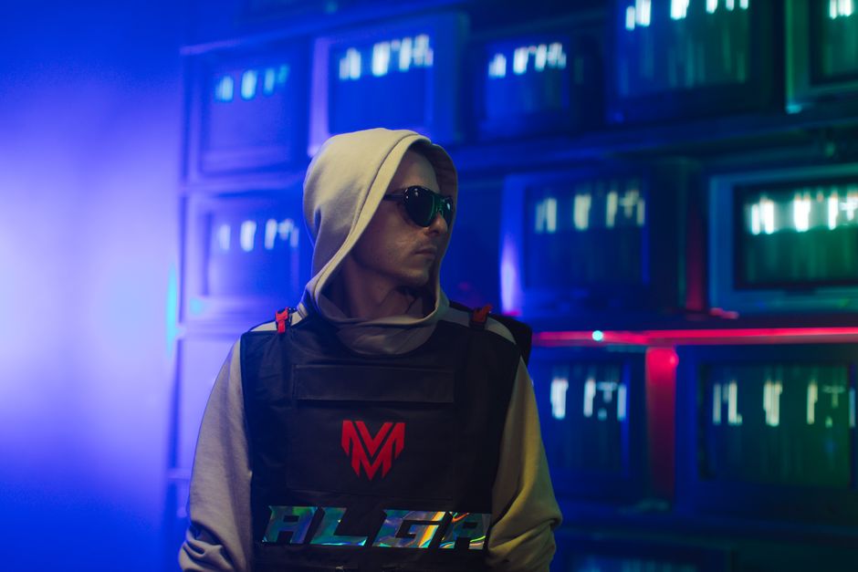 Portrait of a man in a hoodie and sunglasses, with a neon-lit background of screens.