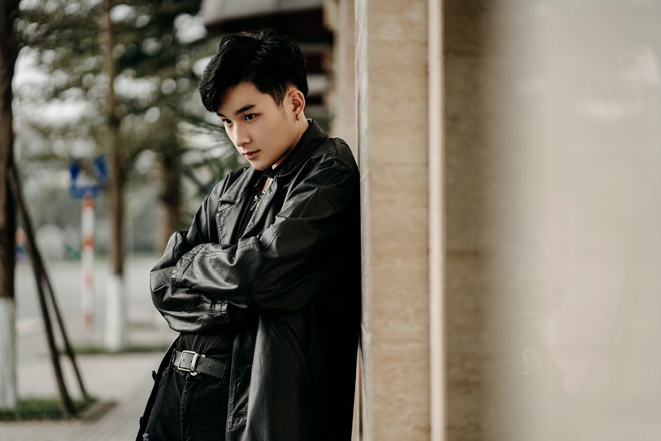 Self assured young Asian male in trendy black leather jacket standing with arms folded and looking away on street