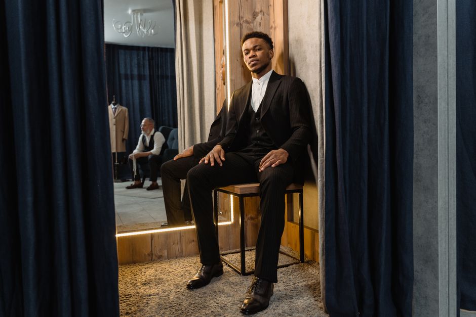 A stylish man in a suit sits in a tailor shop fitting room, embodying elegance and sophistication.
