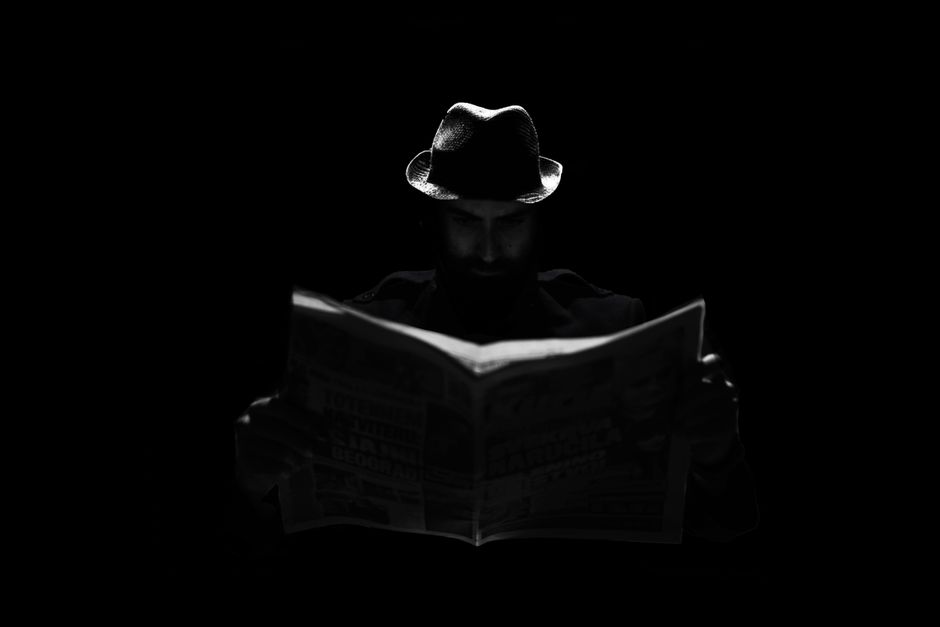 A mysterious figure in a fedora engrossed in reading a newspaper under dramatic lighting.