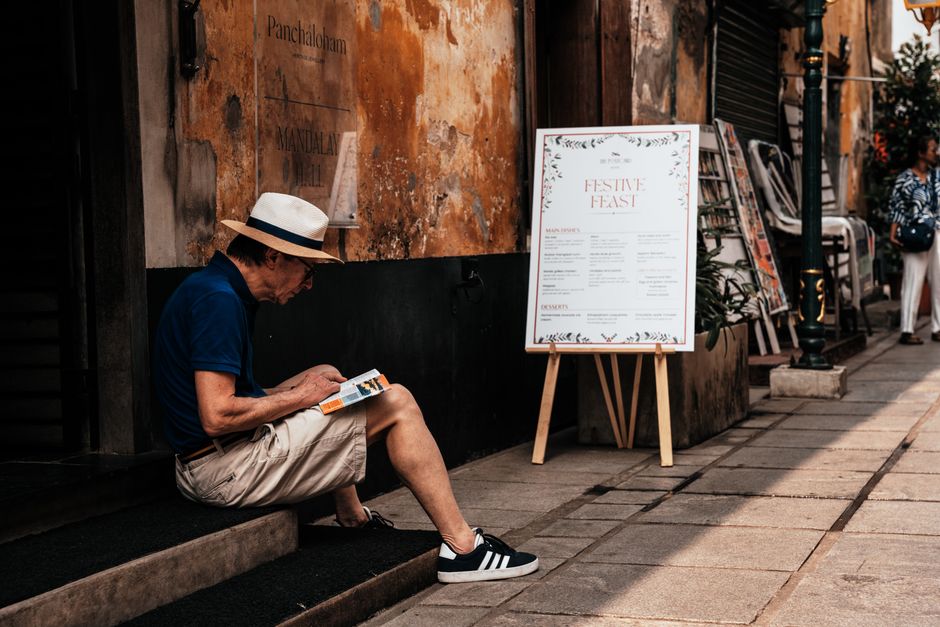 Relaxed man in summer attire reading on a street near a festive menu sign.