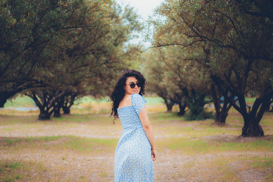Woman in sunglasses and blue dress walking through a serene tree pathway in summer.