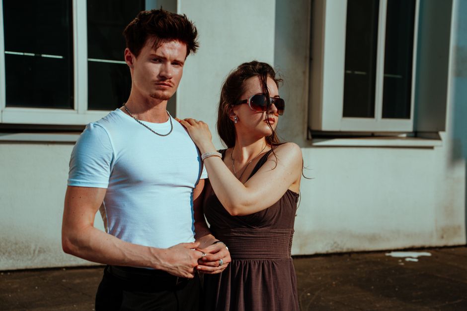 A fashionable couple stands confidently outside a building, styled with sunglasses.