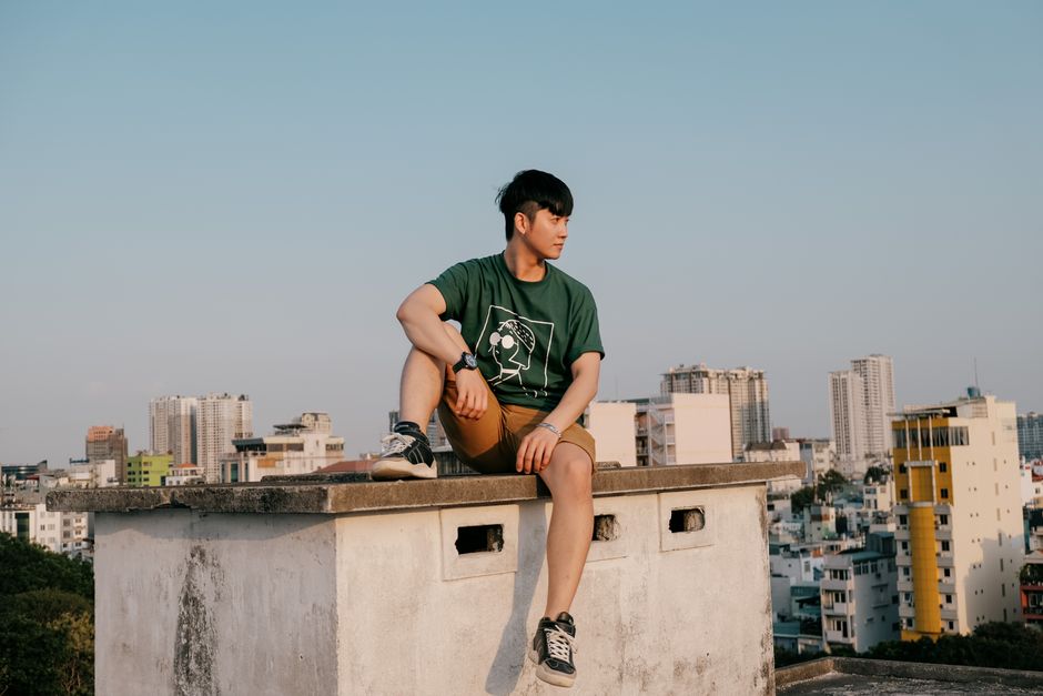 Teenager in casual attire sitting on a rooftop with Ho Chi Minh City skyline in the background.