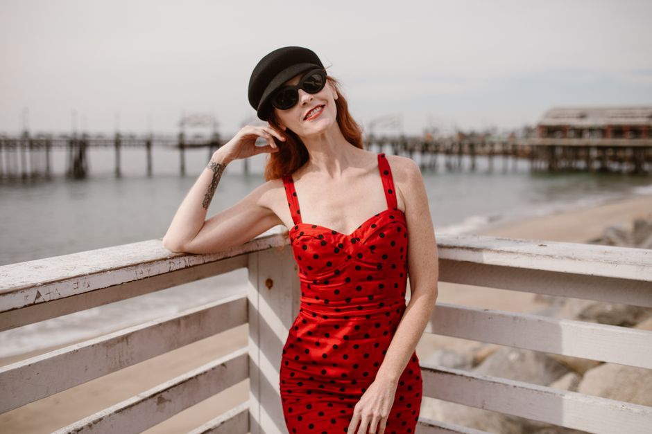 Woman in a red polka dot dress posing by a seaside railing on a sunny day.