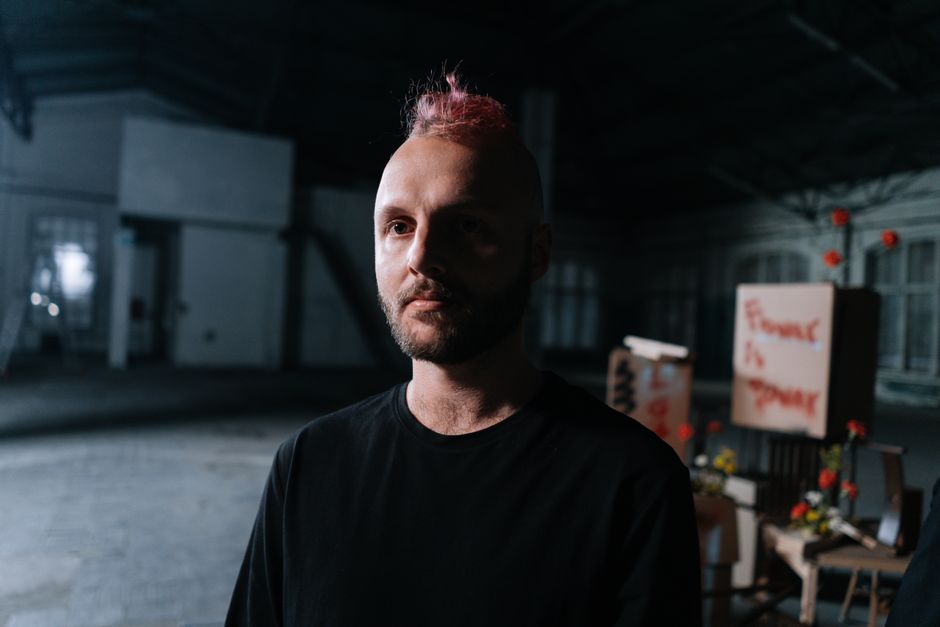 A man with pink dyed hair stands indoors, surrounded by artistic decor, evoking a dramatic mood.