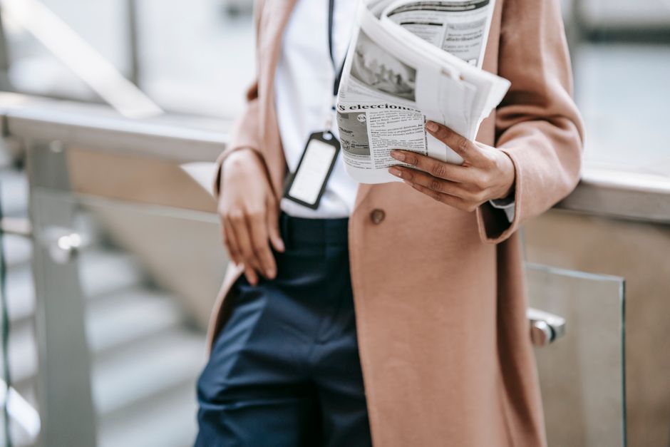 Crop anonymous female entrepreneur in trendy brown coat with badge leaning on fence and reading newspaper